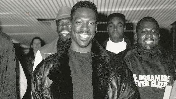 Eddie Murphy in a fur trimmed leather jacket surrounded by people at the airport