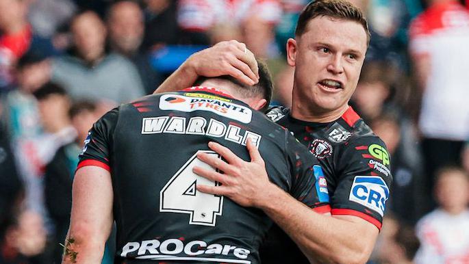 Wigan Warriors player Jai Field smiles and embraces team-mate Jake Wardle after Wardle had scored a try, both players kitted out in black.