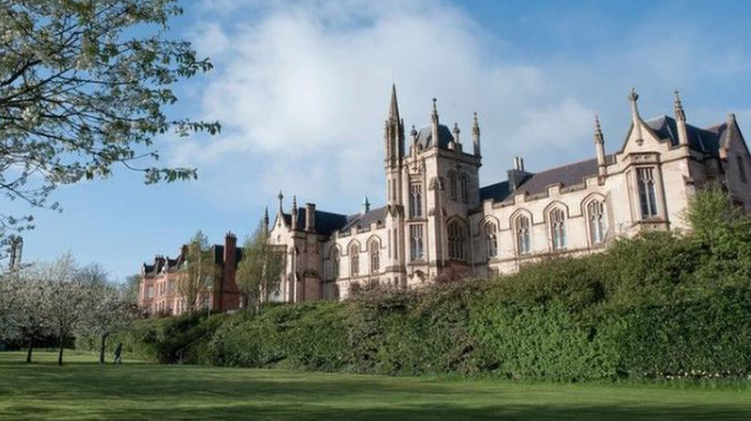 Shows a large university building with spires in front of trees and shrubbery and against the backdrop of a blue cloudy sky