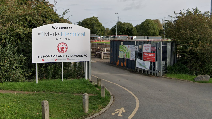 The entrance of Anstey Nomads Football Club with a welcome sign to the left and a football pitch in the distance near trees.