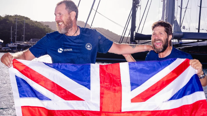 Karl Austen and Ed Shaw smiling at the camera, wearing navy blue t-shirts and are holding a Great Britain flag.