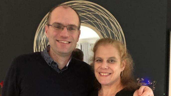 Two people looking and smiling at the camera. A taller man with short dark hair and glasses standing next to a woman with ginger hair tied up.