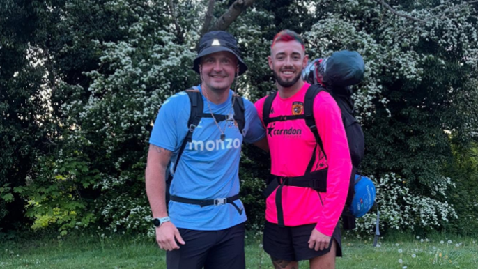 Two men standing in a garden wearing backpacks. The man on the left is wearing a blue football shirt and a bucket hat and the man on the right is wearing a pink long-sleeve top and has pink and blue hair. They have their arm around each other and are smiling. 