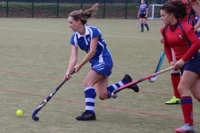 Claerwen running with her hockey stick stretched in front of her next to a yellow hockey ball. She is wearing a blue hockey kit. To the right is a player wearing a red hockey kit with her hockey stick in front of her.