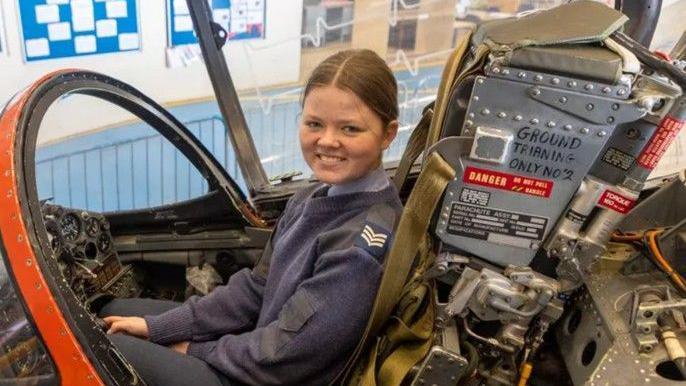 Holly sits in the cockpit of a plane. She looks to the camera and smiles. She wears a navy blue cadet uniform.