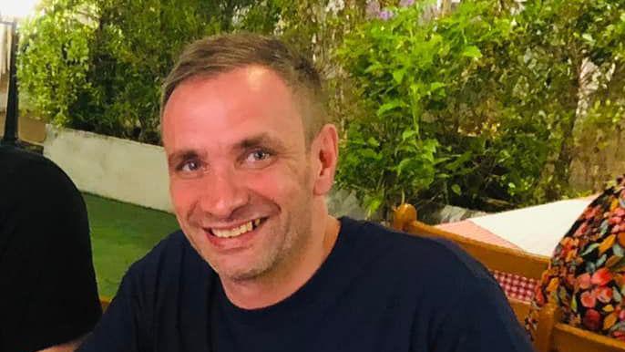 A man with short brown hair and a black t-shirt smiles at the camera