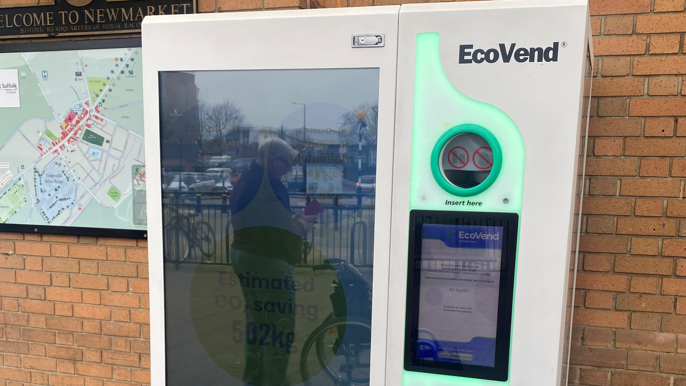 A white EcoVend reverse-vending machine is installed against a brick wall near an entrance. The machine features a large panel on the left and a touchscreen display on the right, illuminated with green LED accents. Above the screen, there are two circular slots for inserting bottles and cans. Visible text on the machine includes 'EcoVend,' 'Insert here,' and 'Swap your empties for exciting rewards.'