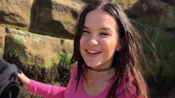 Holli, a dark-haired girl with a black choker and pink top smiling in front of a stone wall.
