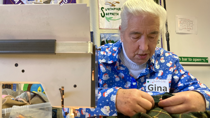 Gina wears a blue Christmas shirt and is concentrating on pushing a needle and thread through material - on the left is the arm of a sewing machine