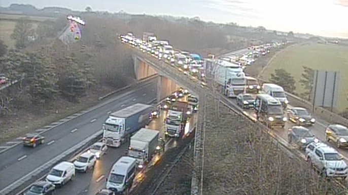 A traffic camera shows the A14 and A11 running over the top with long queues of vehicles. The majority of the vehicles have their headlights on as they wait in the queue.