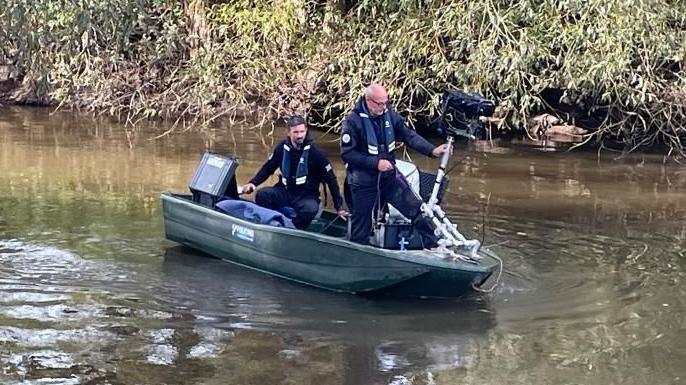 Two men in dark clothing holding search equipment are in a small boat on a murky looking river