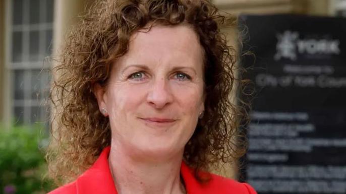 Claire Douglas, a woman with brown curly hair and a red jacket.