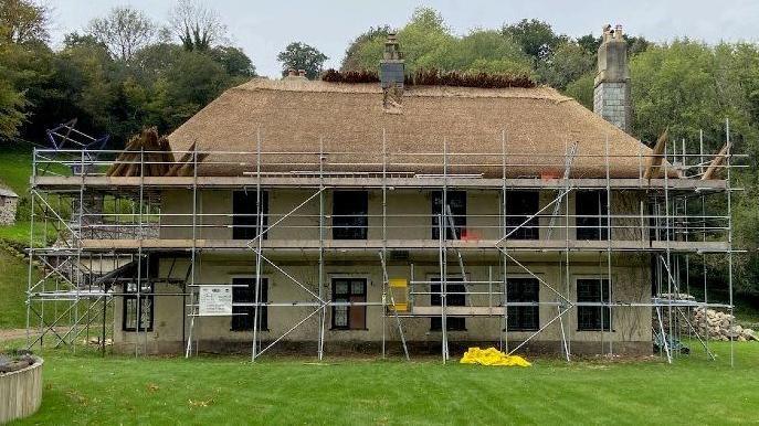 A big house scaffolding up around the whole property. The house has six windows on the ground floor with five on the second floor. Its surrounded by grass with a big fully thatched roof. 