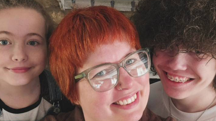 Imogen (centre), Gwen (left) and Reuben (right). All are looking at the camera and smiling. Imogen has auburn hair and a nose piercing, she is wearing glasses.