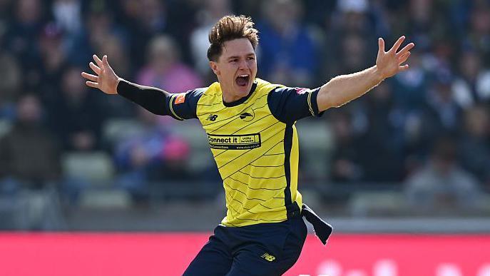 Sonny Baker stands on the pitch with his arms stretched out wide looking over his right shoulder in claim of a wicket for Hampshire