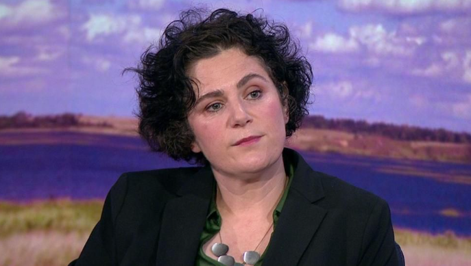 Shows a woman with black hair and a black jacket green top and necklace looking at a camera
