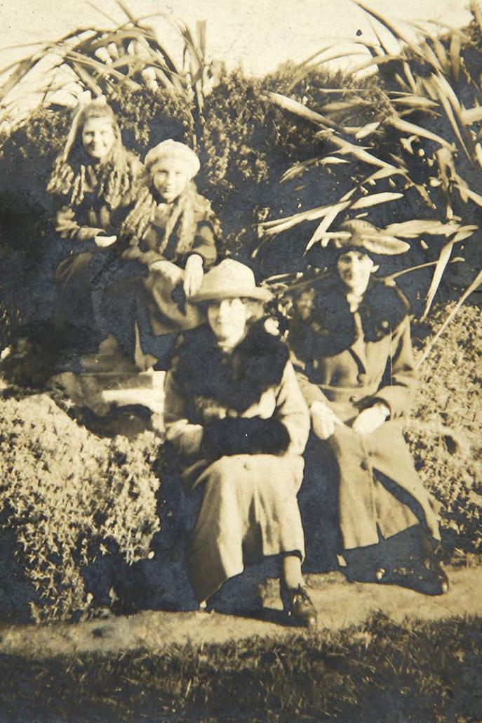 A black and white photo of a group of people on an outing sat on what appears to be a hedge next to a conifer. Two older woman have long coats on and hats and two young girls in the background also have scarves on and coats.