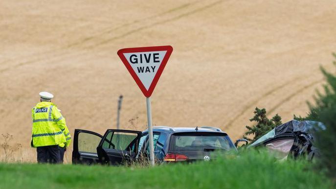 A police officer stands at a junction in the road between green grass and a brown crop field. Next to him there is a give way sign, a black Volkswagen Golf and the wreckage of another car.