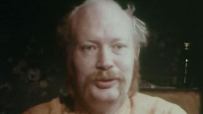 Sam Green. He is balding with ginger sideburns and moustache that grows down to his chin at the sides.