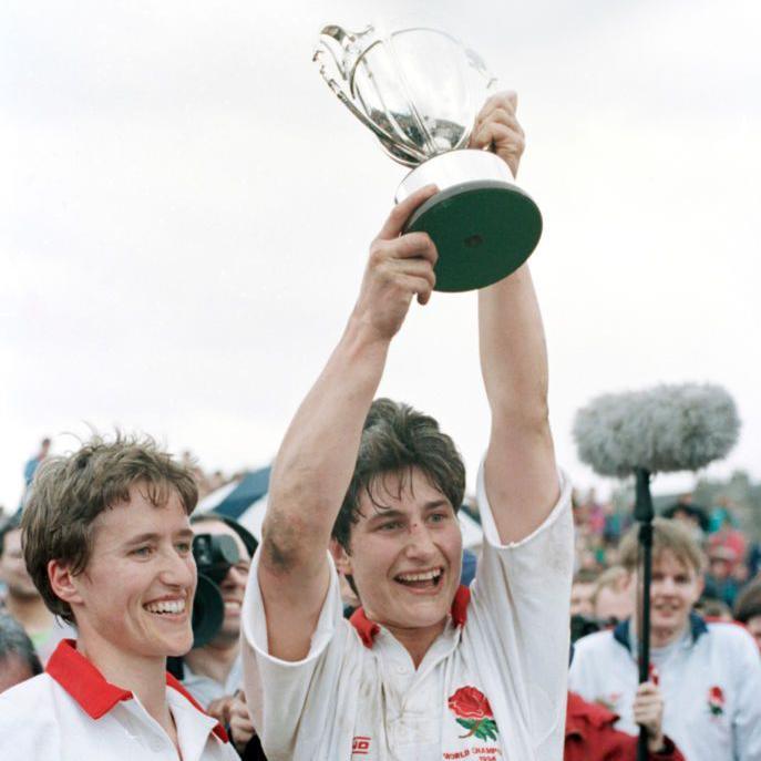 A woman with short brown hair smiles broadly as her teammate lifts a silver trophy. They are wearing white rugby shirts with red trim. A crowd stands behind them, including a film crew. 