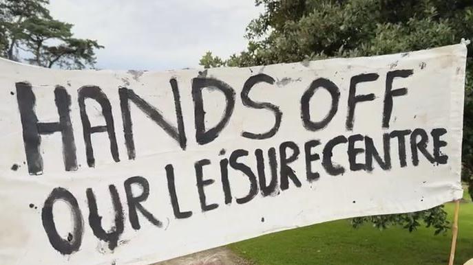 A handmade protest banner held up outside the Ards and North Down Council meeting.   It reads: "Hands Off Our Leisure Centre" in black paint across a white background. 