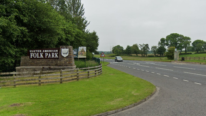 A Google Maps screenshot of the Ulster American Folk Park sign, which is situated beside a main road. There is a car driving towards the sign, with a number of fields on the right hand side of the image and large trees planted around the sign.