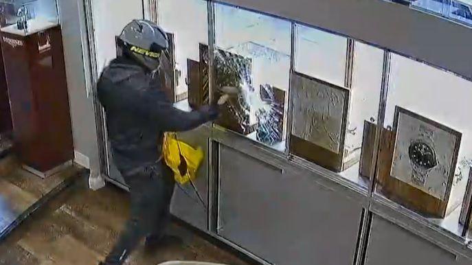 A CCTV image of a man wearing dark clothing and a motorbike helmet smashing glass cabinets in a jewellery shop with a hammer while carrying a yellow bag