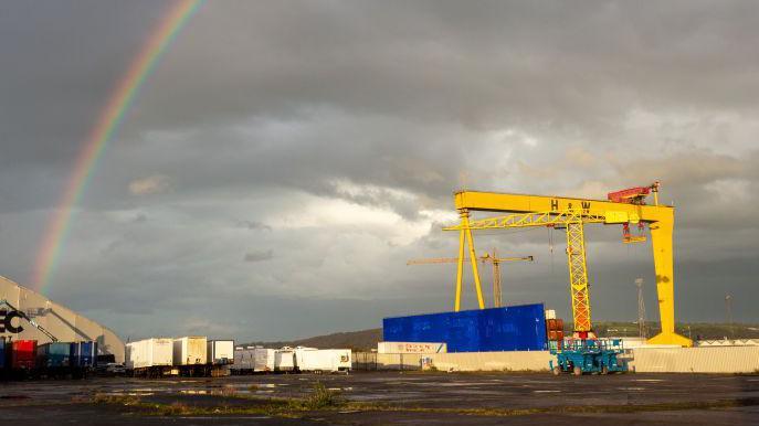 A yellow crane with H&W on it. The sky in the background is grey. There is a rainbow in the sky.