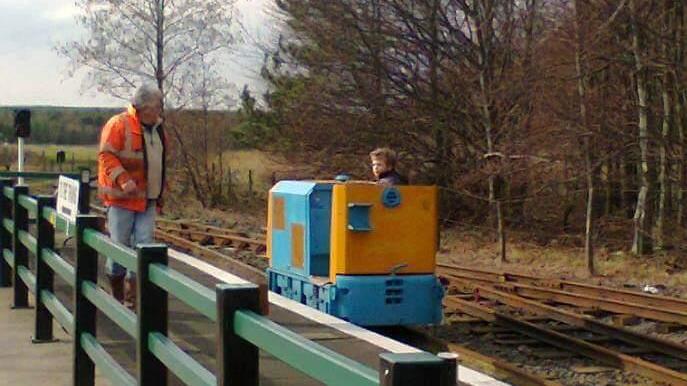 Logan as a young boy in a small blue and yellow train carriage on a railway track. An older man stands on the platform wearing a hi-vis orange jacket, looking on.