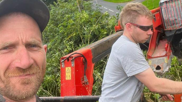 Daniel Graham, wearing a black cap and with a short ginger beard, takes a selfie as Adam Carruthers, with short blond hair, sunglasses and a grey t-shirt, cuts at some timber with a chainsaw.