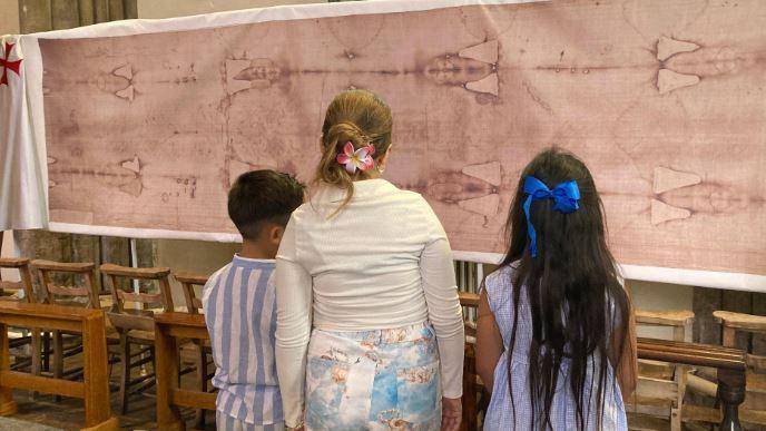 Three children stand in front of the large cloth with a faint image of a man’s body imprinted on it. The children wear light-coloured clothes and two girls have flowers or bows in their hair.
