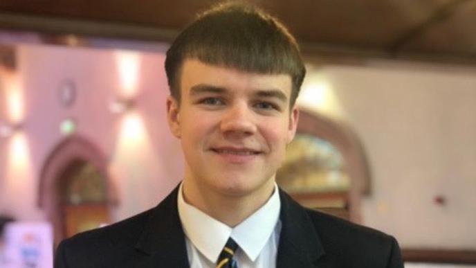 Gerry is in a school uniform. He has short brown hair and is wearing a black school blazer, white shirt and multi-coloured school tie. 