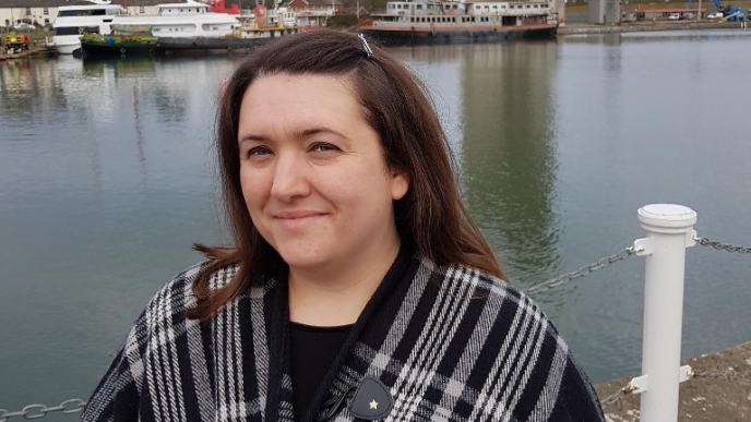 Stroud District Councillor Lindsey Green stood next to water at a docks, with boats in the background. She has long dark hair and a black and white cardigan on.
