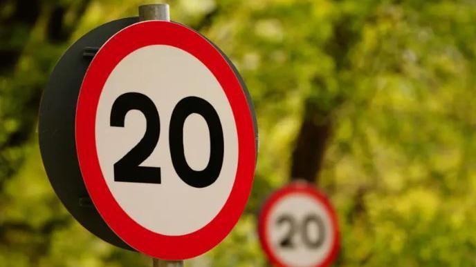 A 20mph limit road sign, with a red ring around it. In the distance is a further road sign carrying the same warning, with blurred trees in the background. 