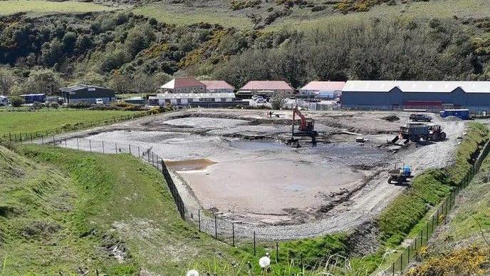 A large area containing silt which is fenced off within a green field. There is a digger and other machinery within the compound, with large industrial units behind.