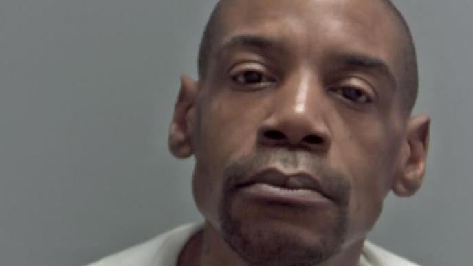 A police custody photo of Derek Campbell, who has tilted his head to the side while staring at the camera. He has a skinhead and black stubble. His is wearing a light grey top.