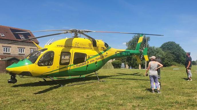 A yellow and green Wiltshire air ambulance helicopter has landed on a patch of grass next to houses, there are three onlookers.