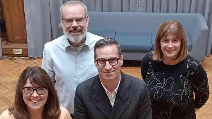 Luton Rising managing director Nick Platts and Head of Commercial Operations Linsey Sweet (back row) with Active Luton Chief Executive Officer Lawrence Kay and Director of Finance and Resources Sue Jones (front), all looking up at a camera, smiling, they are in a room with a blue sofa behind them and standing on a parque floor.
