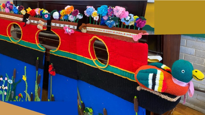A narrow boat, made out of fabric, with a duck on the front, flowers on the top, and the narrowboat is made out of red and yellow wool. 