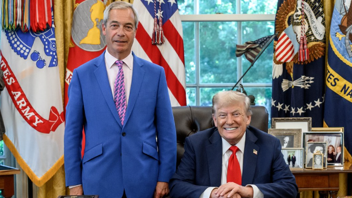 Nigel Farage stands beside a seated Donald Trump in the Oval Office. Farage wears a blue suit with a purple tie, while Trump is in a dark suit with a red tie. They are behind the Resolute Desk, which holds phones, documents, and other items. The room features large windows showing greenery outside, with American and military flags, statues, and framed photos decorating the space.