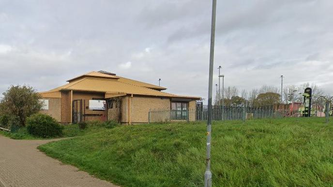 Single-storey light brick building (now known as Kingswood Neighbourhood Centre) with three narrow windows with dark frames to the right of the building. There is a two-stage pitched roof and there are railings from the right side of the building. There is a grass area in front of the building.