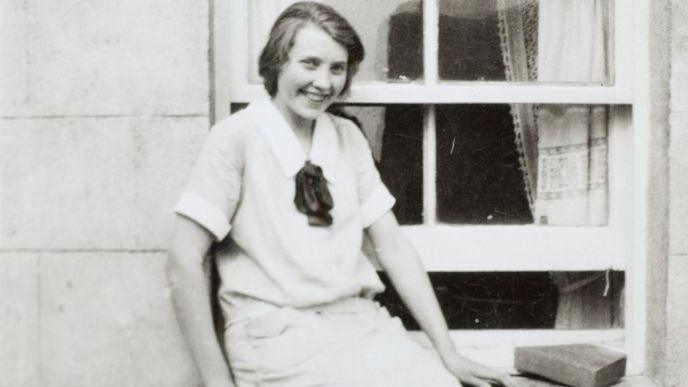 A black and white photo of a smiling young woman, Mary Anne MacLeod, in 1928. She is sitting on a window ledge of a house, and she is wearing a light top with black necktie and a light skirt. Her hair appears to be quite dark and is quite short, parted on the right.