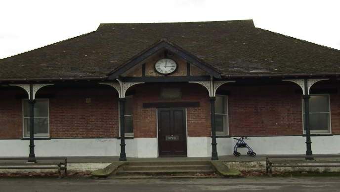 the outside of a long single-storey sports pavilion, with slim sash windows and steps leading up to the door under a covered porch with Victorian-style pillars and a clock above the door