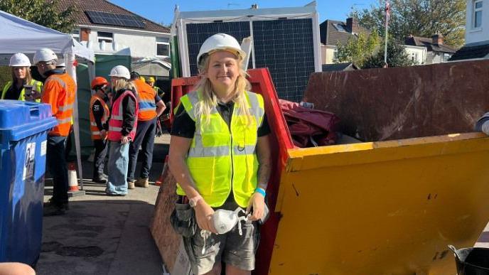 Cat Adelova is wearing a fluorescent yellow vest and a white hard hat. She is standing next to a large yellow skip. There are other tradespeople in florescent vests in the background.