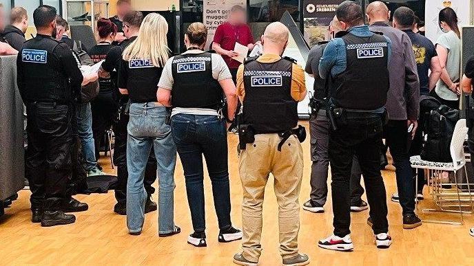 Police officers, some in uniform and some plain-clothed, all wearing stab vests, are briefed in a room with a wooden floor.