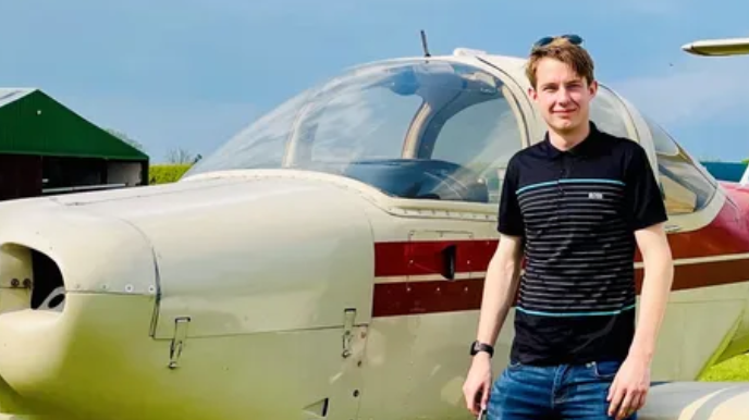 A man in a black polo shirt with white stripes stands in front of a light aircraft, smiling. It is a sunny day.