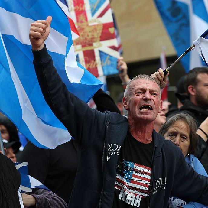 A man raises his fist while standing in front of a group of people waving flags, including saltires and a union flag.