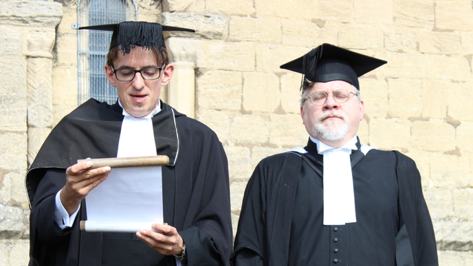 Two men stood outside the light stone bricked chapel. They are wearing black robes with a white collar and university graduation hats on their heads. The man on the left is reading from a scroll. 