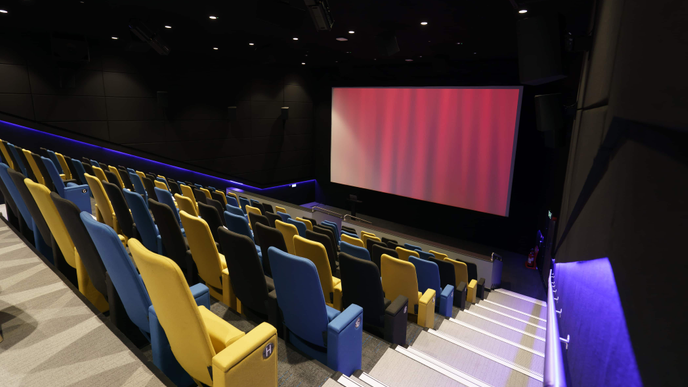 Cinema auditorium with rows of yellow, black, and blue seats, shown from the back. The screen is lit up in red. The staircase parapet is lit up in blue. 