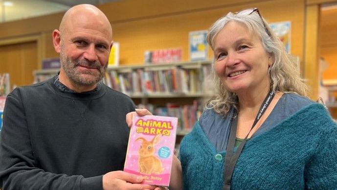 A man and a woman holding a copy of a book while standing in front of a bookshelf. They are both smiling. The man is bald and wearing a dark grey sweater. The woman has a knitted blue cardigan on over a blue top. The book is pink and has a rabbit on its cover.